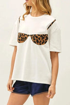 A woman modeling a white short-sleeve t-shirt with a leopard-print bra graphic on the front, paired with navy shorts and brown boat shoes, against a plain white studio background.