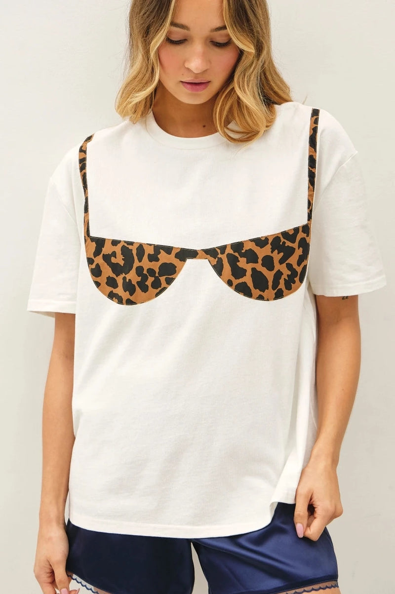 A woman wearing a white short-sleeve t-shirt with a novelty leopard-print bra graphic on the chest, paired with navy blue satin shorts, against a plain light gray background.