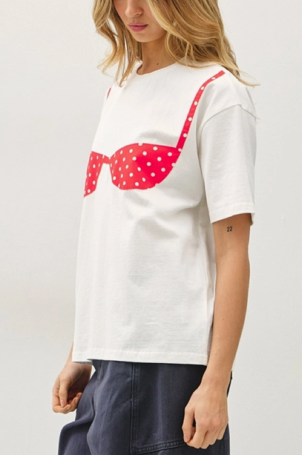 A woman wearing a white short-sleeve t-shirt with a red polka-dot bra graphic across the chest, paired with dark blue pants, against a light gray background.