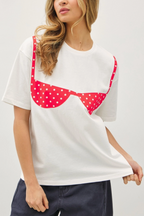 A woman wearing a white short-sleeve t-shirt with a red and white polka dot bra graphic across the chest, paired with dark blue jeans against a light gray background.