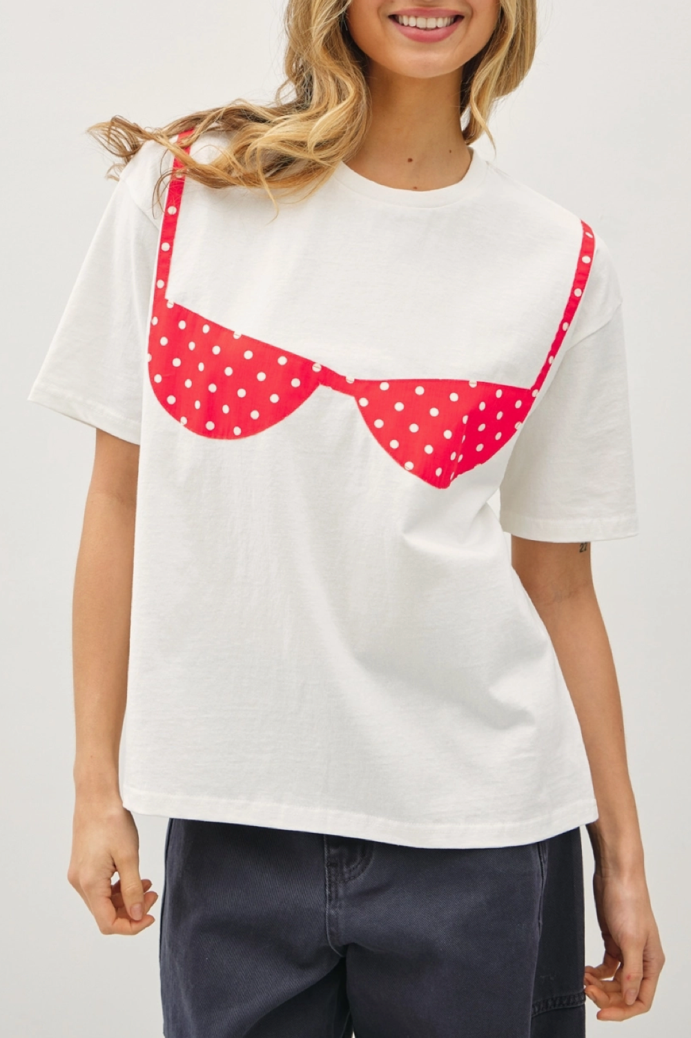 A white short-sleeve t-shirt featuring a novelty graphic that looks like a red bra with white polka dots and straps, modeled by a woman in a studio setting.