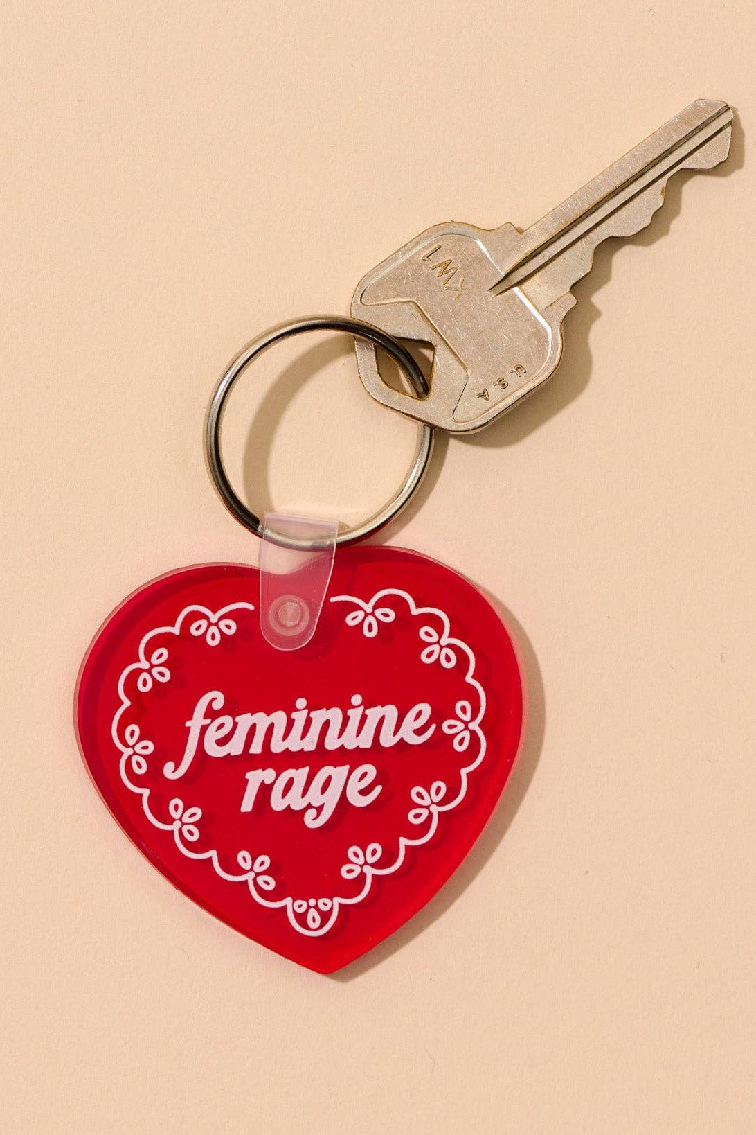 Red heart-shaped keychain with 'feminine rage' text on a beige background