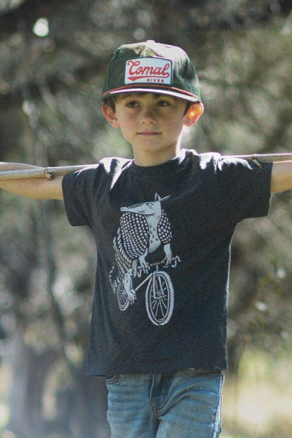 Child wearing a cap and t-shirt with a graphic design, standing outdoors with trees in the background