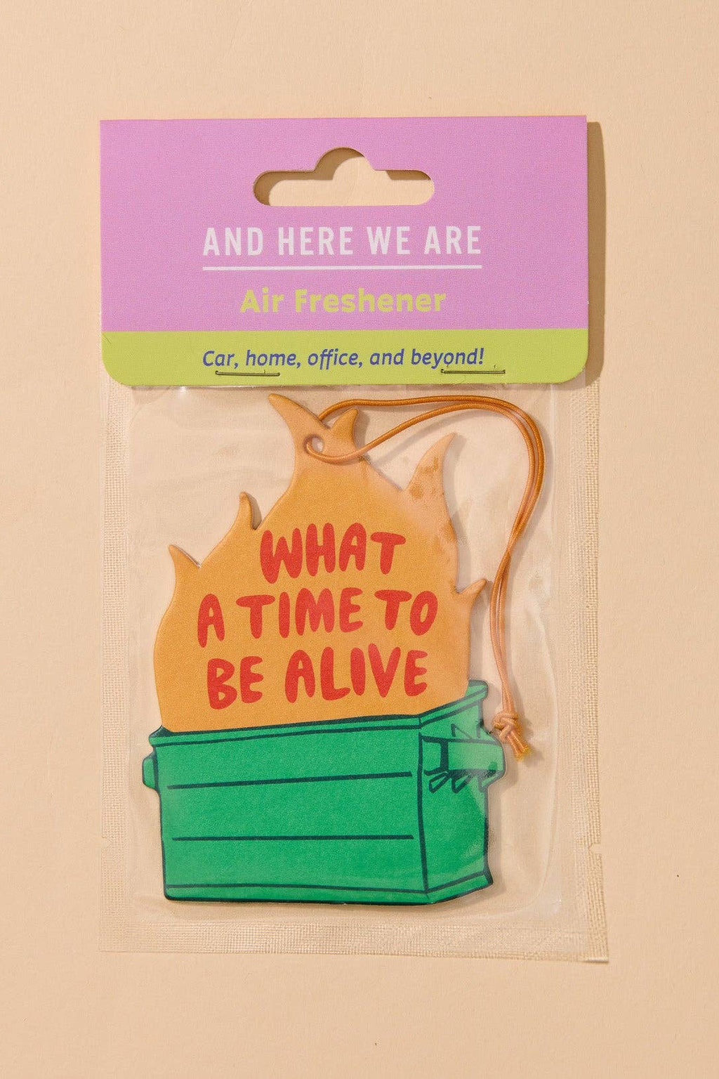 A packaged novelty air freshener shaped like a green storage box with a yellow flame-shaped top that reads “WHAT A TIME TO BE ALIVE.” The item is attached to a pink and green card header that says “AND HERE WE ARE Air Freshener.