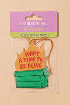 A packaged novelty air freshener shaped like a green storage box with a yellow flame-shaped top that reads “WHAT A TIME TO BE ALIVE.” The item is attached to a pink and green card header that says “AND HERE WE ARE Air Freshener.