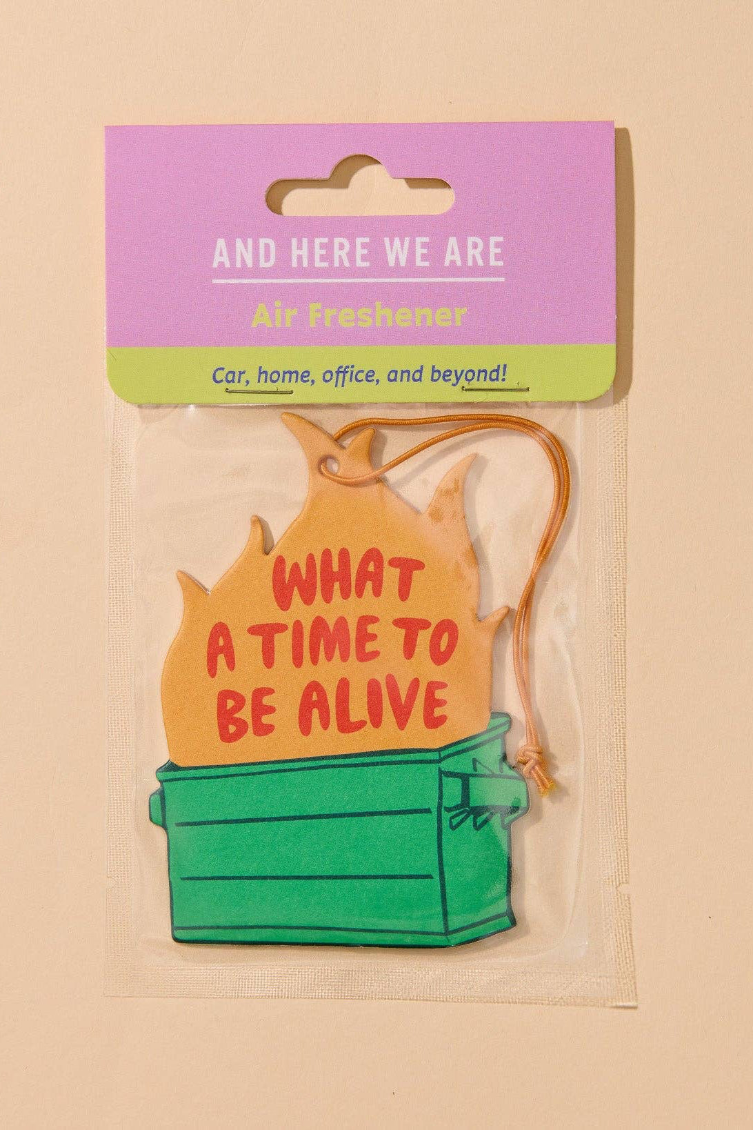 A packaged novelty air freshener shaped like a green storage box with a yellow flame-shaped top that reads “WHAT A TIME TO BE ALIVE.” The item is attached to a pink and green card header that says “AND HERE WE ARE Air Freshener.