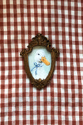 A small framed duck portrait with an ornate gold-toned frame, featuring a white duck with an orange beak and a blue bow, displayed on a red and white checkered fabric background.