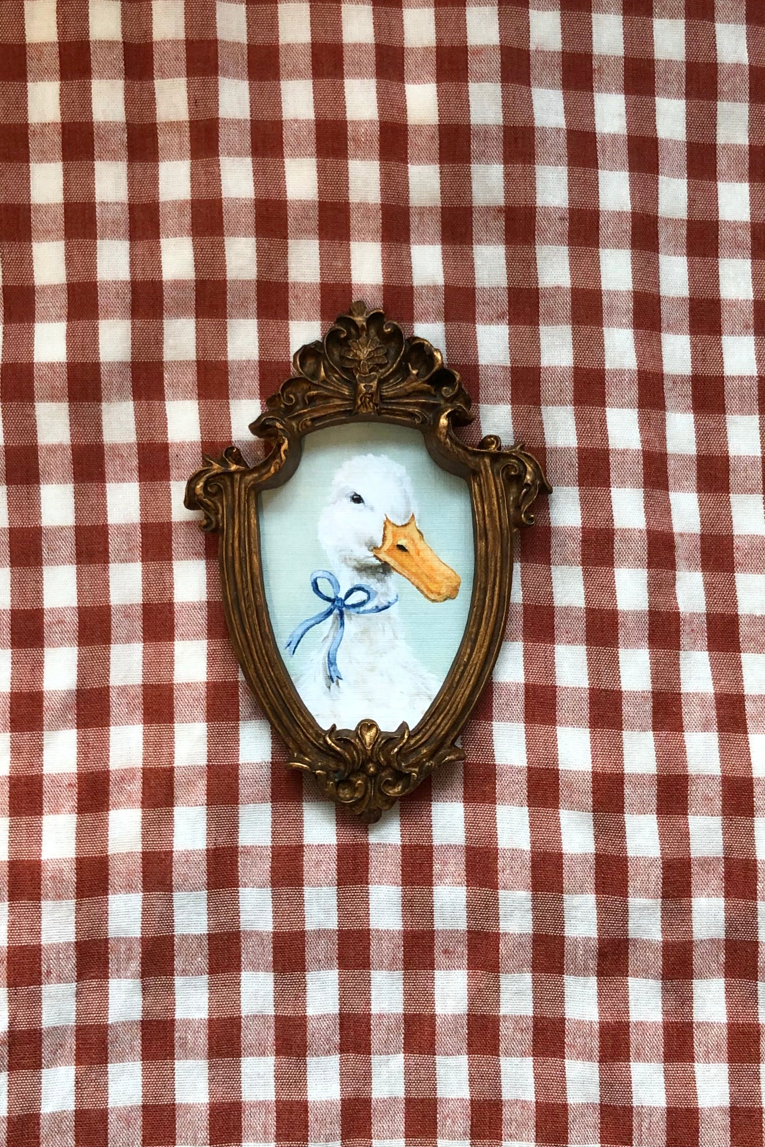A small framed duck portrait with an ornate gold-toned frame, featuring a white duck with an orange beak and a blue bow, displayed on a red and white checkered fabric background.