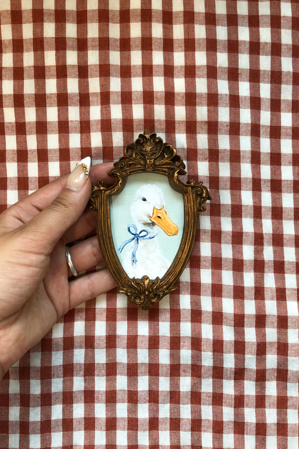A hand holding a small decorative oval frame featuring a painted duck with a blue bow and yellow beak on a white background, set against a red and white checkered fabric backdrop.