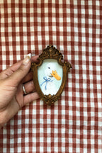 A hand holding a small decorative oval frame featuring a painted duck with a blue bow and yellow beak on a white background, set against a red and white checkered fabric backdrop.