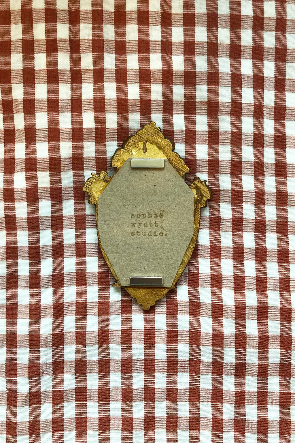 A vintage gold-tone oval pin or badge with a beige backing, placed on a red and white checkered fabric background. The backing card has embossed text including 'SOPHIE WYATT STUDIO'.