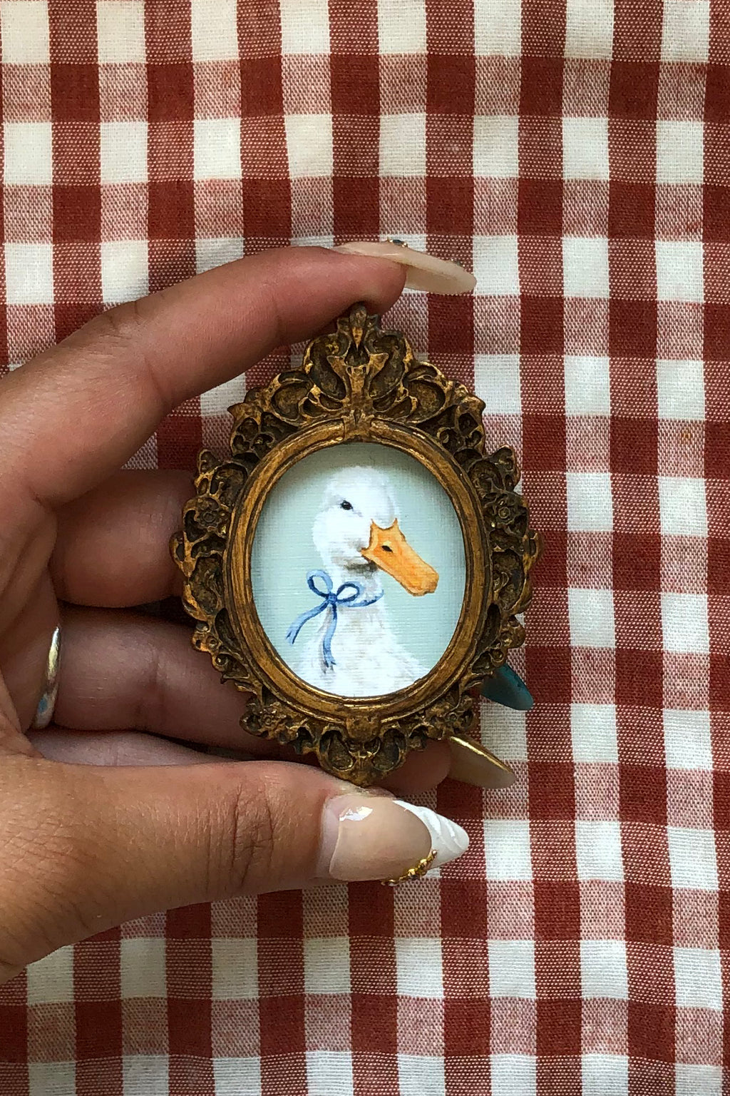 A hand holding a small ornate gold-framed portrait of a white duck with an orange beak and a blue bow, set against a red and white checkered fabric background.