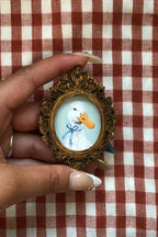 A hand holding a small ornate gold-framed portrait of a white duck with an orange beak and a blue bow, set against a red and white checkered fabric background.