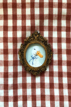 A small oval framed portrait of a white duck with a blue bow and orange beak, set in an ornate gold-toned frame and placed on a red-and-white checkered fabric background.