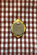 A decorative oval pin or badge with a distressed gold border and a gray center stamped with 'sophia wyeth studio', placed on a red and white checkered fabric background.