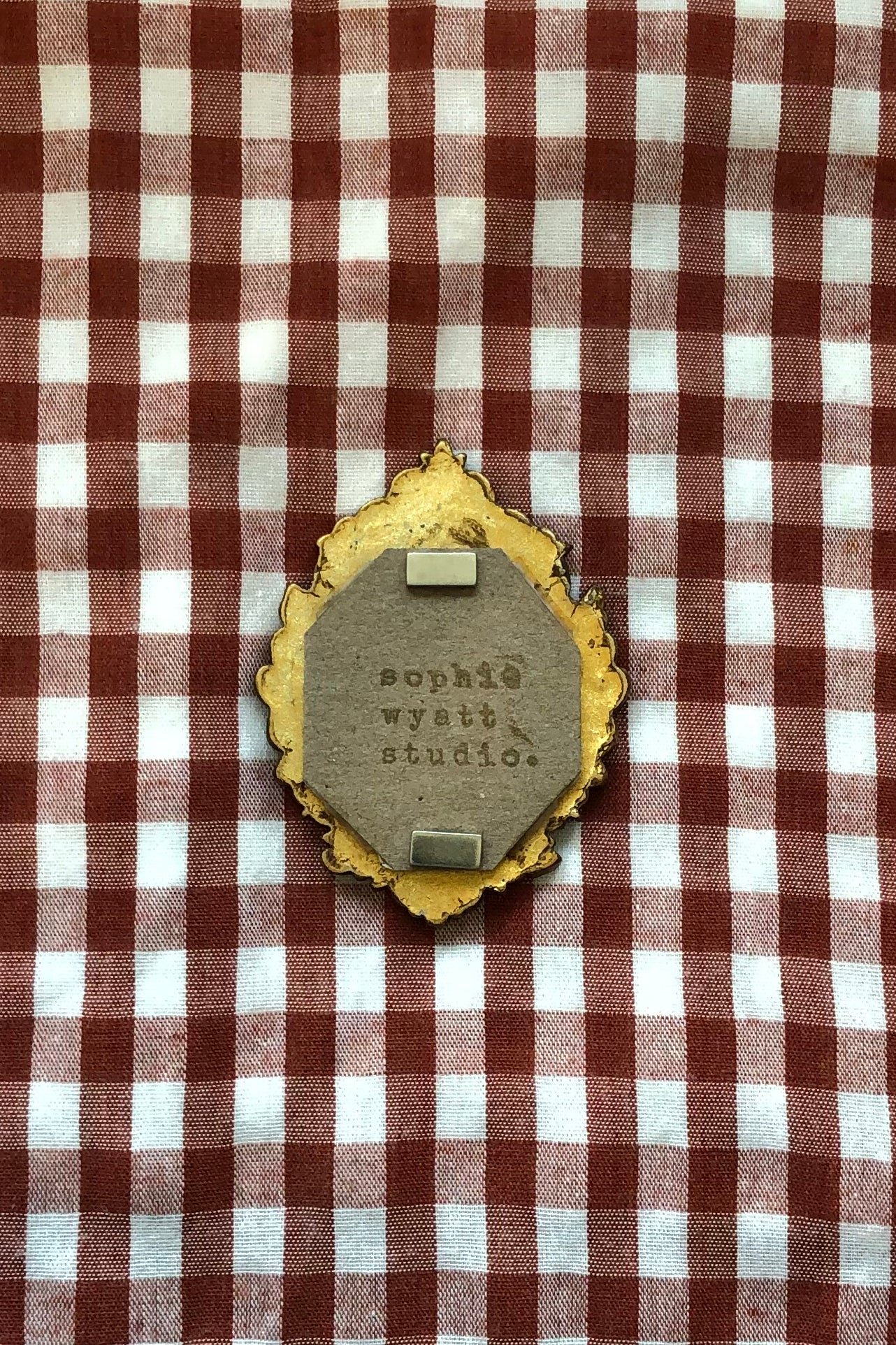 A decorative oval pin or badge with a distressed gold border and a gray center stamped with 'sophia wyeth studio', placed on a red and white checkered fabric background.