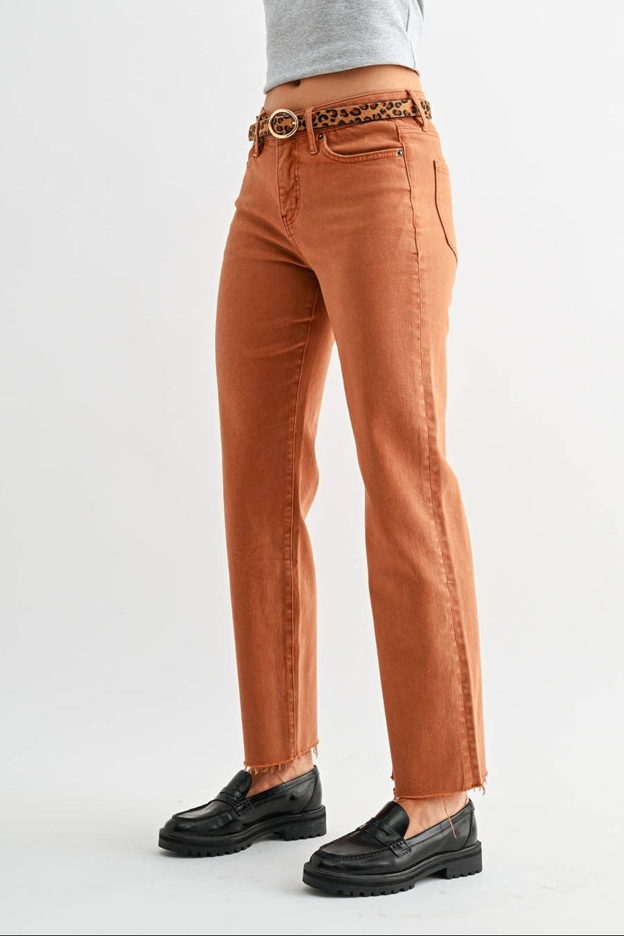 A woman modeling a pair of rust-orange straight-leg jeans with a brown leather and patterned chain belt, paired with a light gray crop top and black loafers, against a plain light gray studio background.