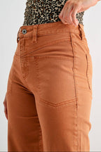 A close-up of a model wearing rust-orange denim jeans, showing the side pocket, belt loop, and button. The model is also wearing a leopard-print top, set against a plain light gray studio background.