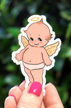 Hand holding a small angel sticker against a green leafy background