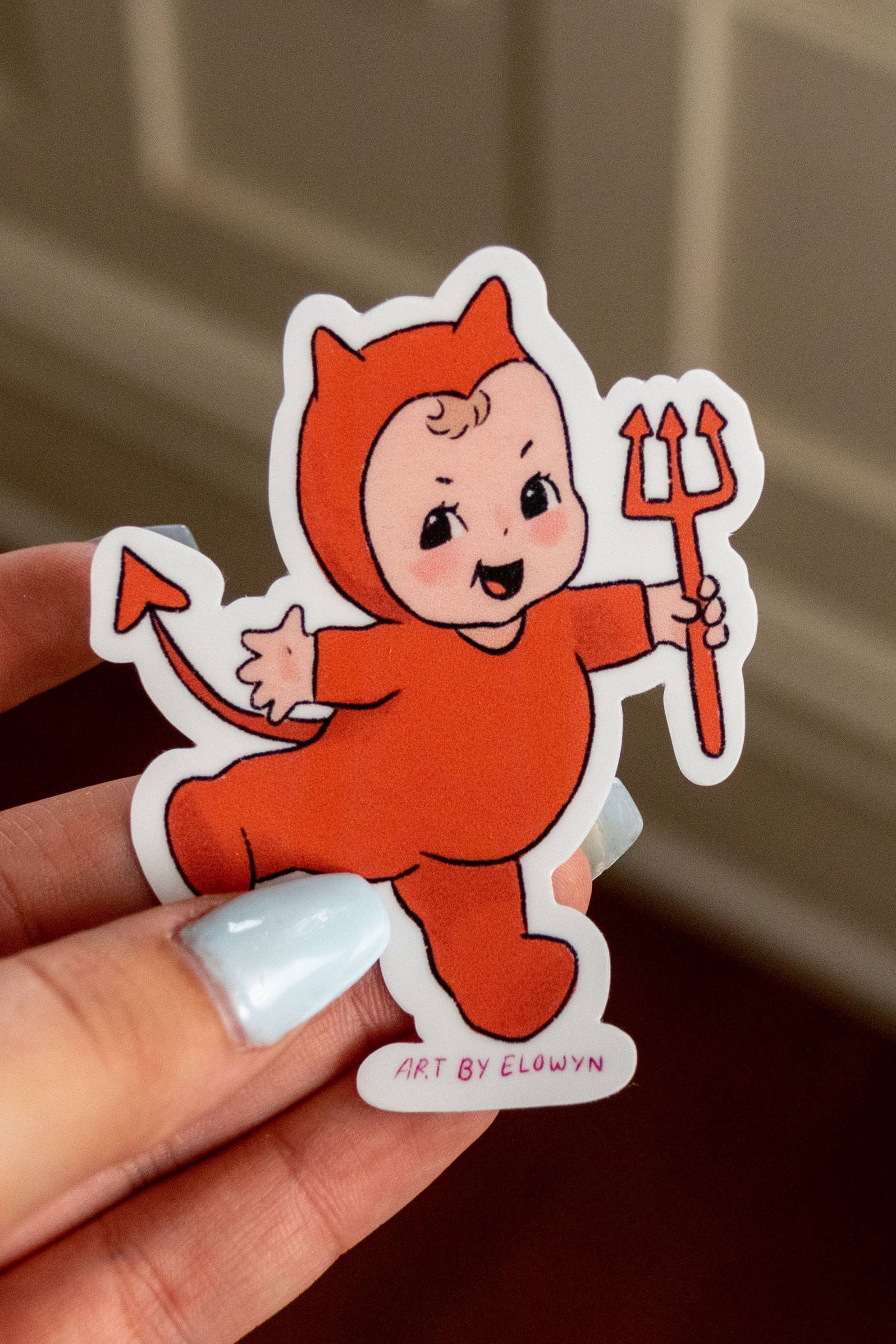 Hand holding a sticker of a small child in a devil costume against a neutral background