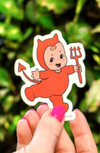 Hand holding a sticker of a cartoon character with devil horns against a green leafy background