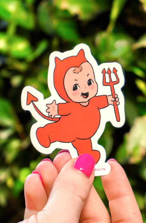 Hand holding a sticker of a cartoon character with devil horns against a green leafy background