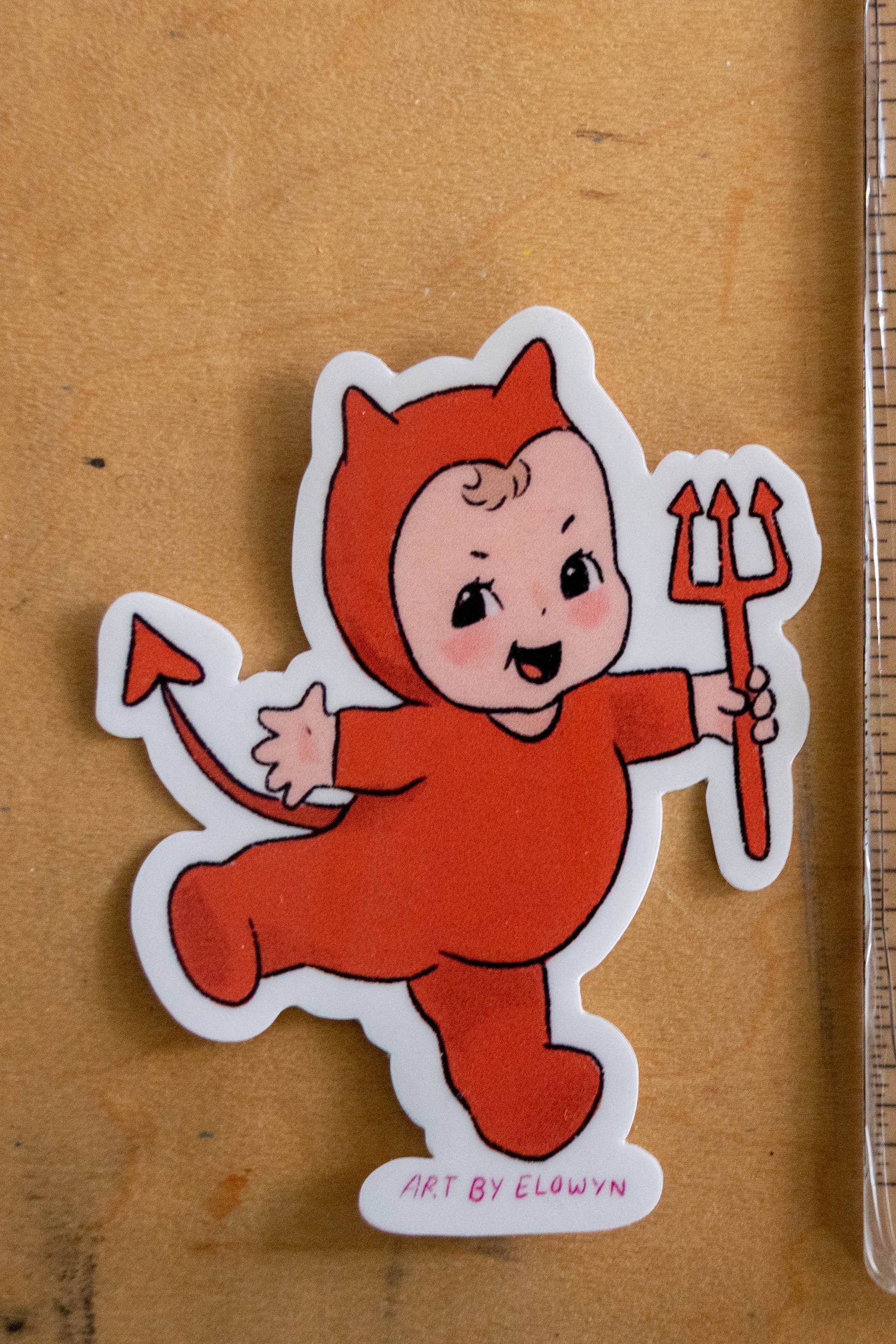 Sticker of a child in a red devil costume next to a ruler on a wooden surface
