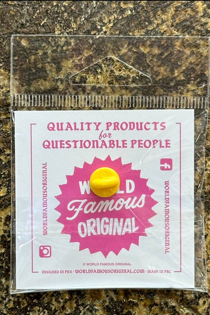 A small round yellow pin badge with a pink and white paper backing card, sealed in a clear plastic bag and placed on a mottled brown and black stone surface. The card reads 'QUALITY PRODUCTS for QUESTIONABLE PEOPLE' and features a pink starburst with the words 'WORLD Famous ORIGINAL'.