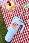 White tumbler with a cake design on a red and white checkered tablecloth with a dessert in the background.