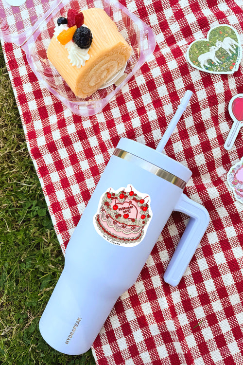 White tumbler with a cake design on a red and white checkered tablecloth with a dessert in the background.