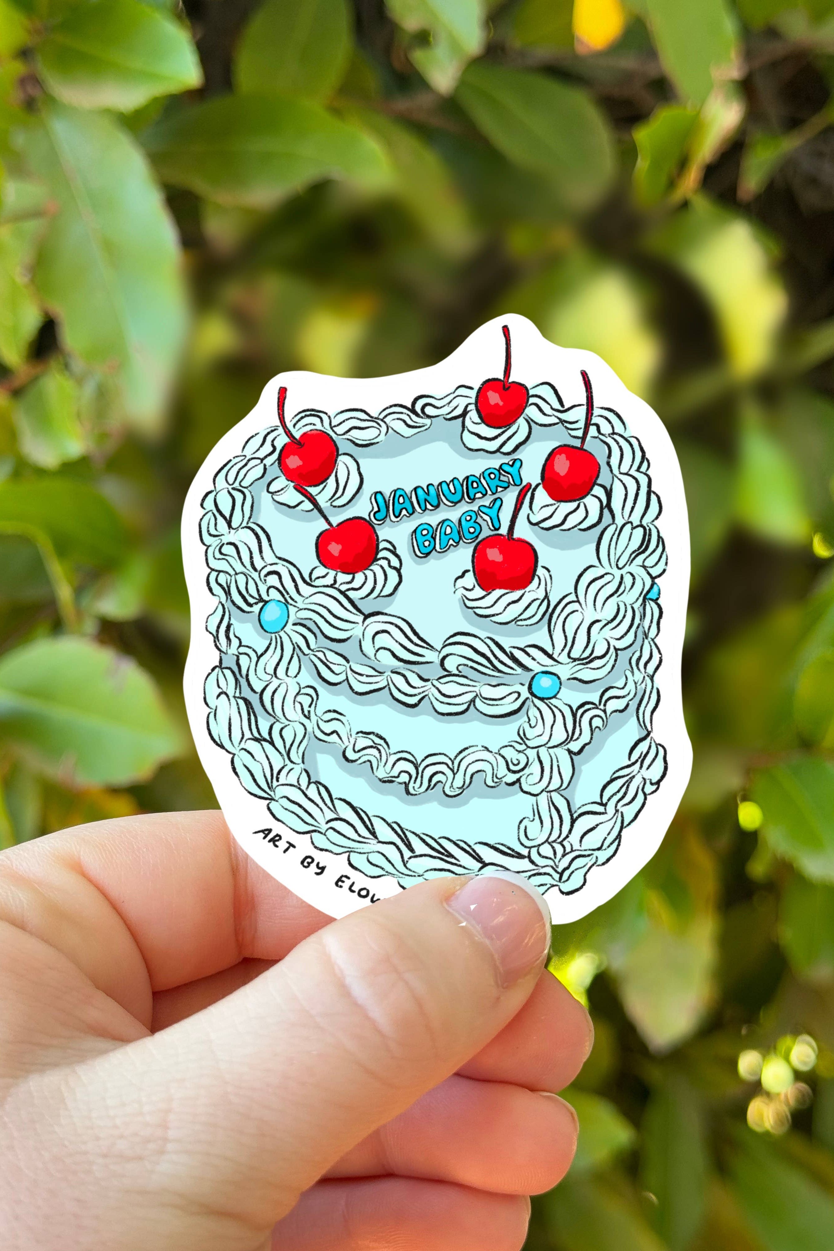 Hand holding a sticker with cherries and text against a green leafy background