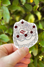Hand holding a sticker with 'July Baby' design against a green leafy background