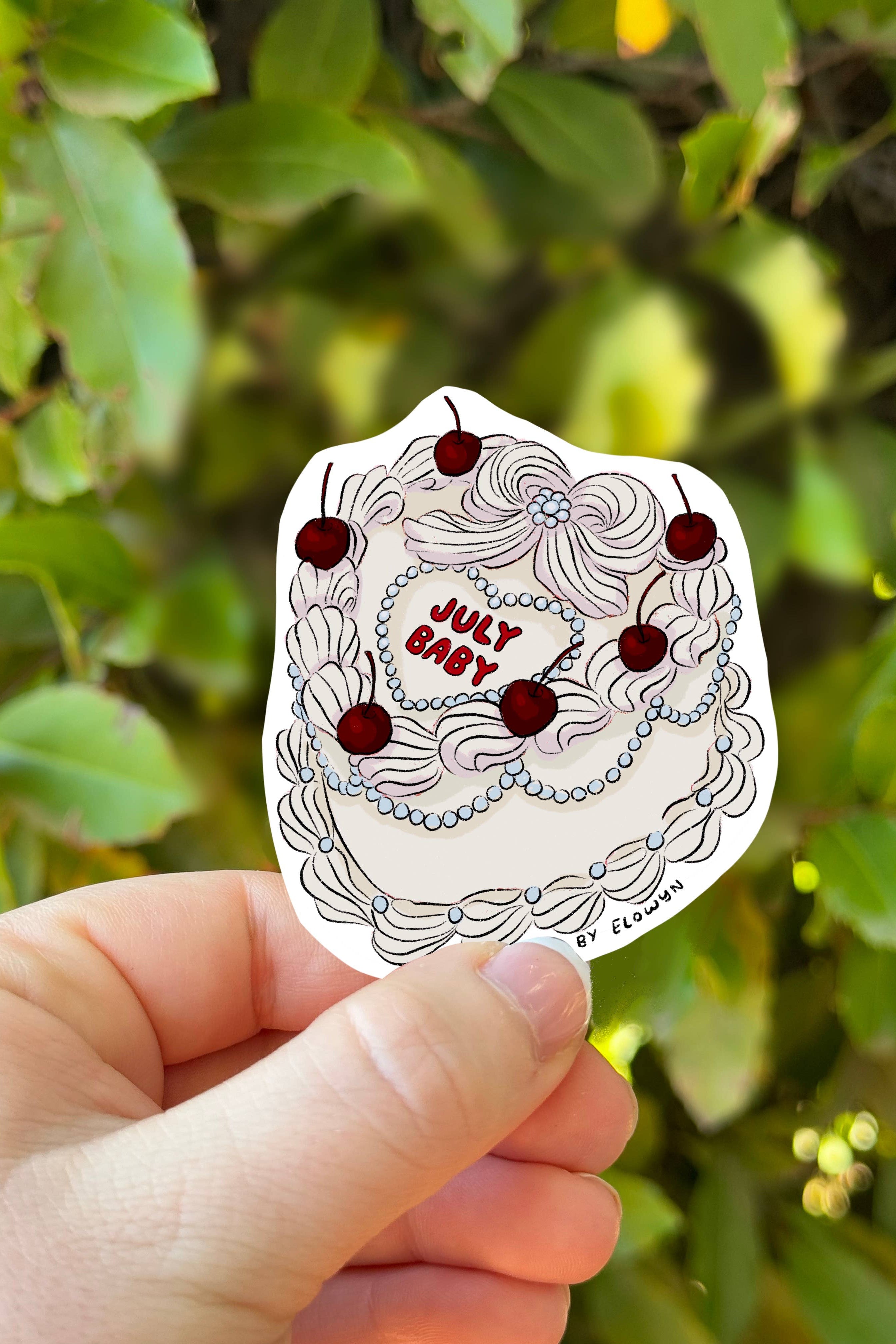 Hand holding a sticker with 'July Baby' design against a green leafy background