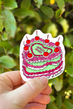 Hand holding a colorful cake-shaped sticker with red cherries against a green leafy background