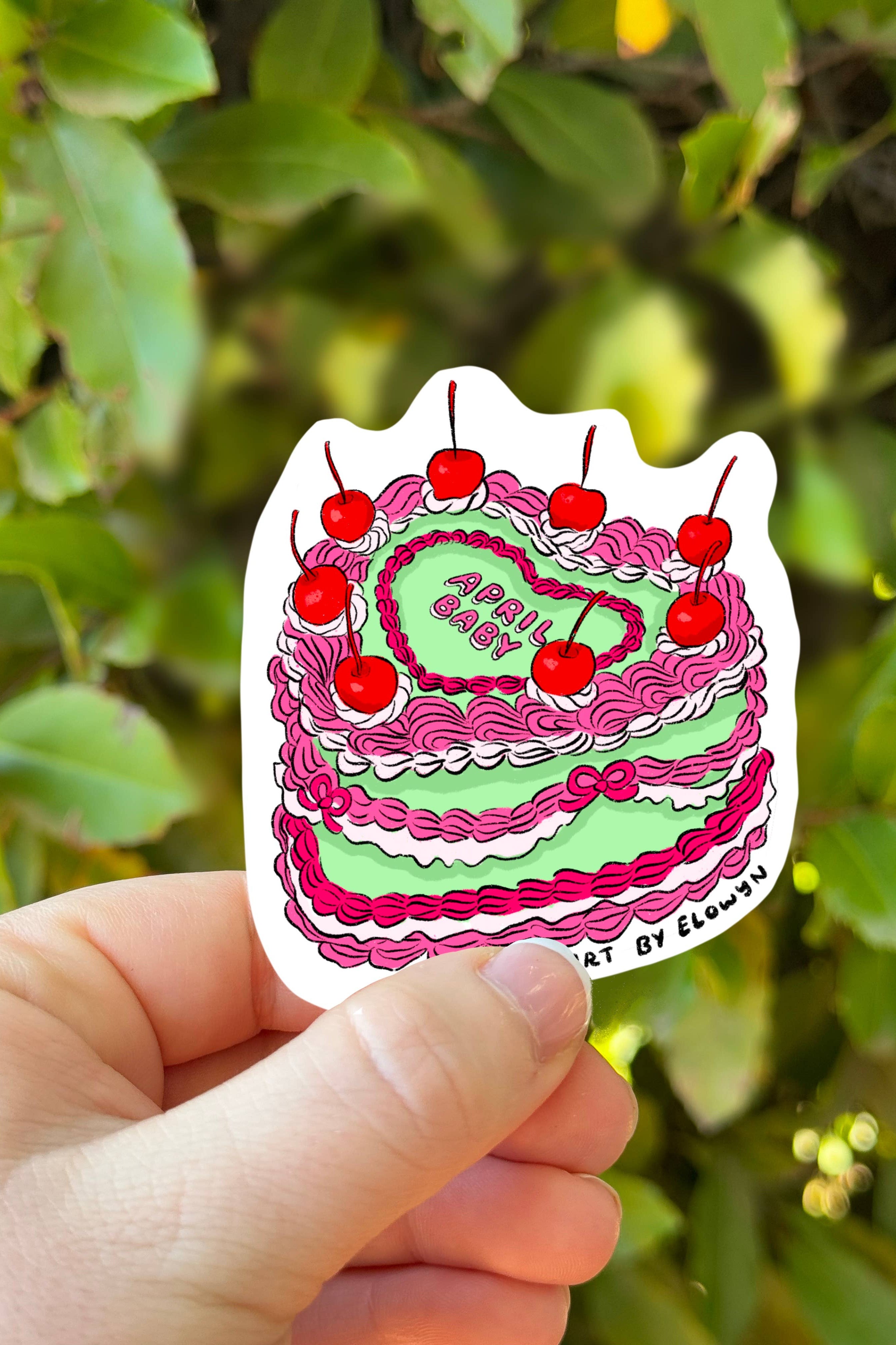 Hand holding a colorful cake-shaped sticker with red cherries against a green leafy background