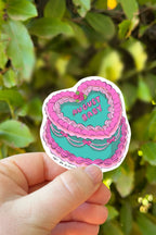 Hand holding a sticker with a cake design and 'August Baby' text against a green leafy background.
