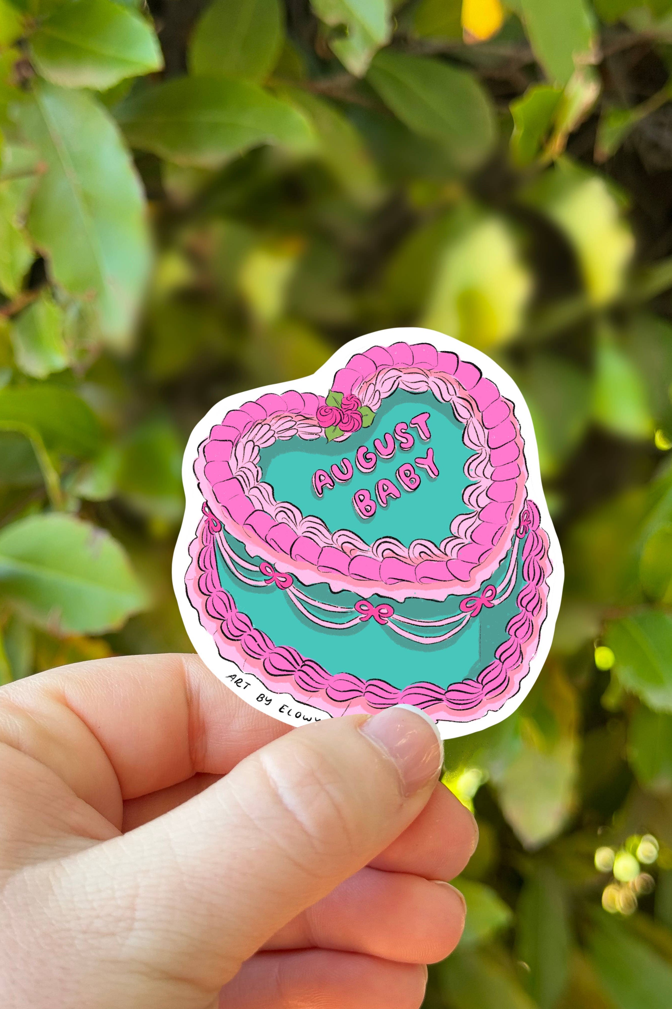 Hand holding a sticker with a cake design and 'August Baby' text against a green leafy background.
