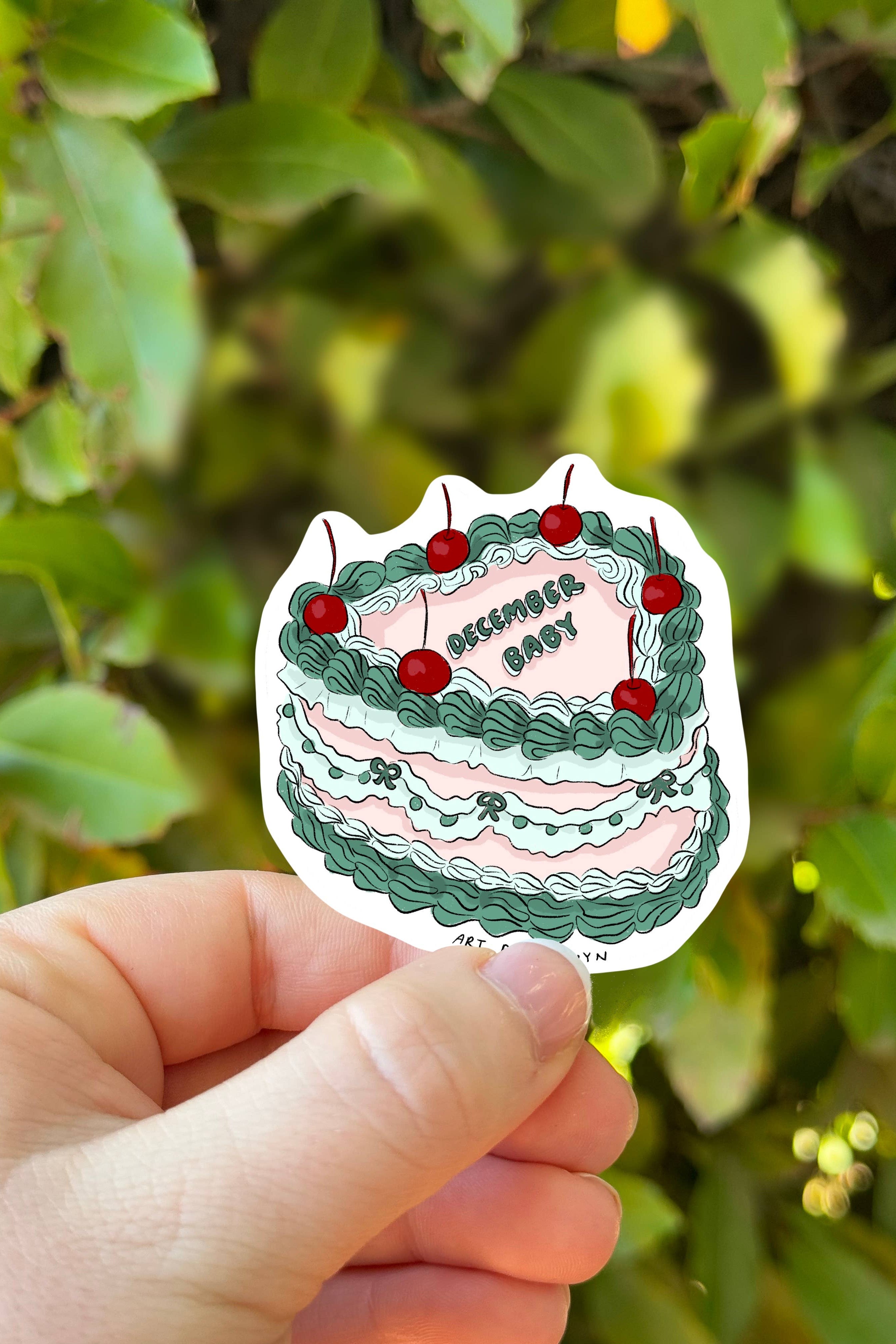 Hand holding a cake-shaped sticker with green and pink frosting against a leafy green background