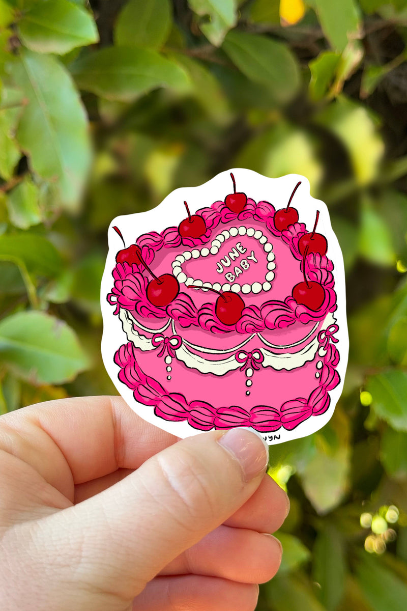 Hand holding a pink cake-shaped sticker with red cherries against a green leafy background