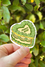 Hand holding a sticker with green and yellow design against a leafy background