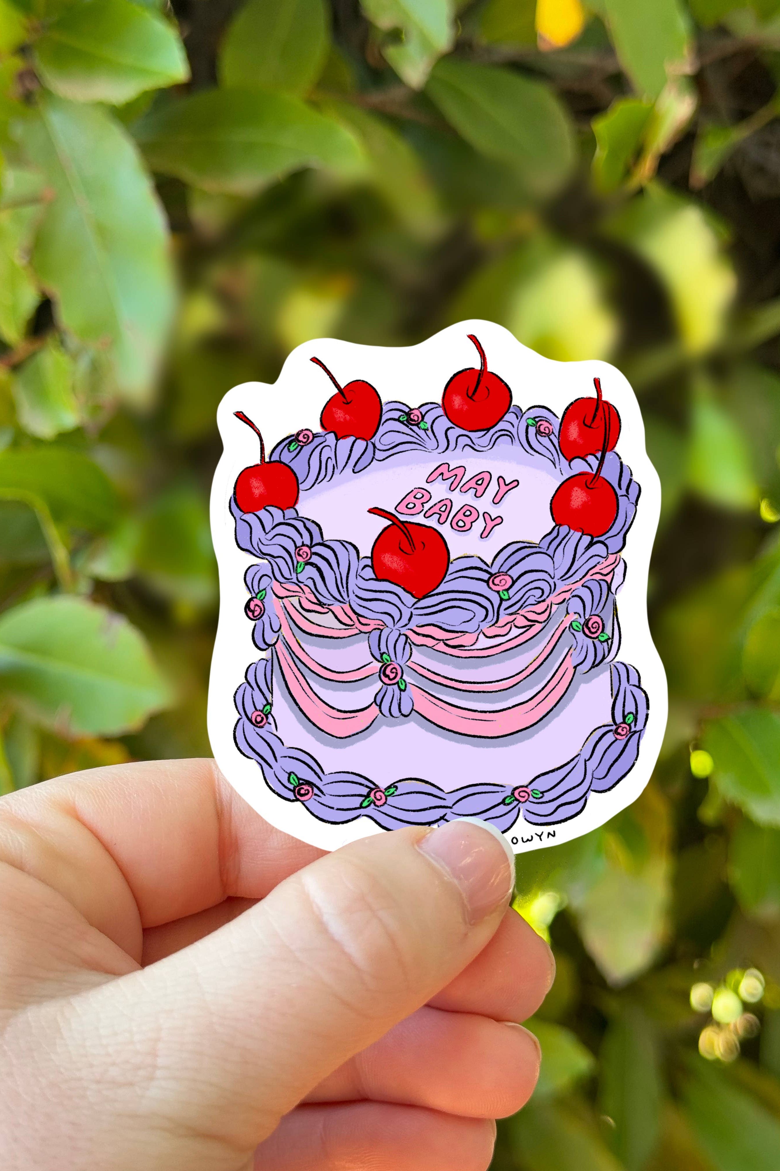 Sticker with cherries and text held by a hand against a green leafy background