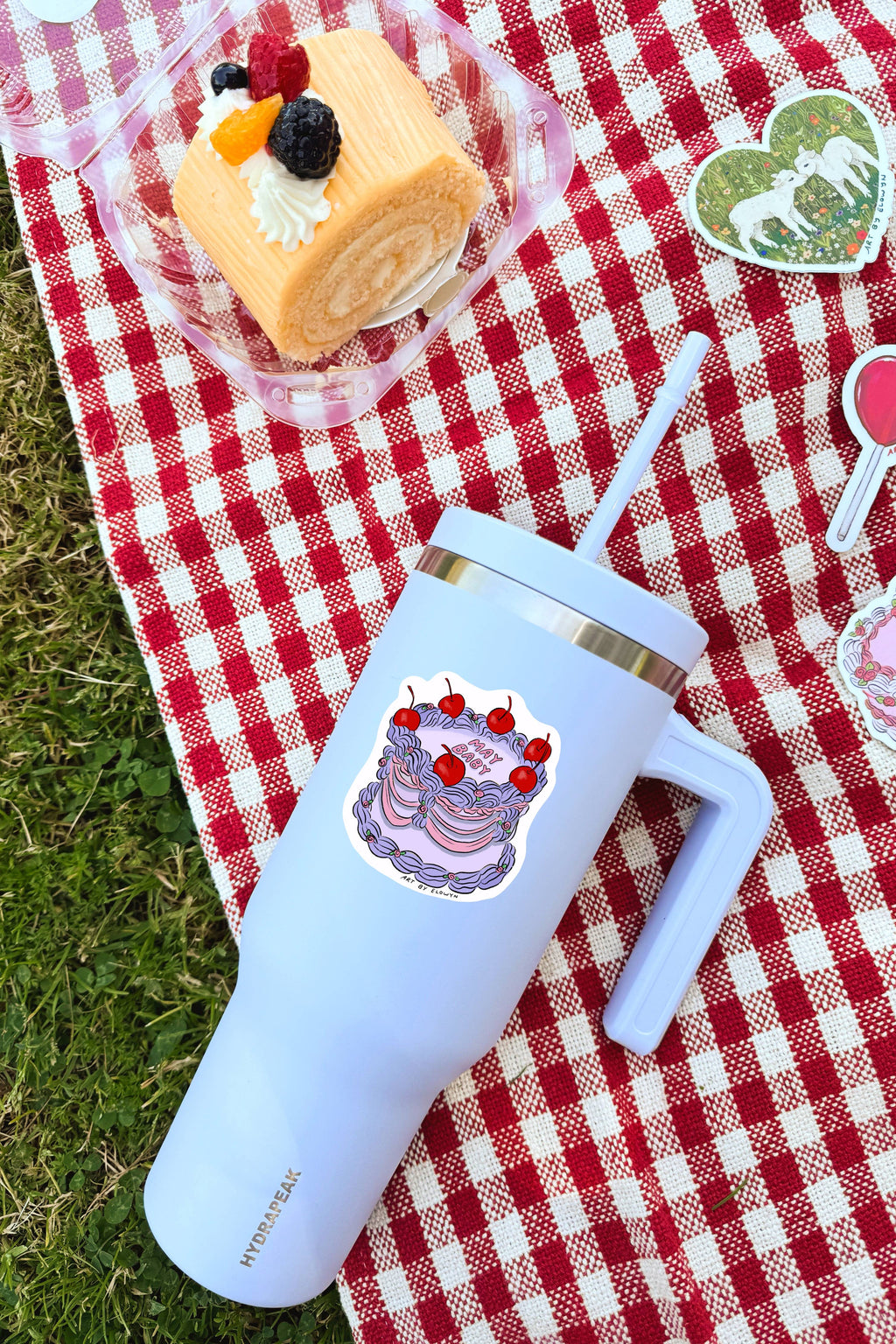 Light blue insulated tumbler with cake design on a red and white checkered tablecloth with a dessert in the background.
