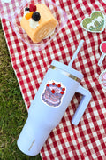 Light blue insulated tumbler with cake design on a red and white checkered tablecloth with a dessert in the background.