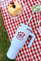 Light blue insulated tumbler with cake design on a red and white checkered tablecloth with a dessert in the background.
