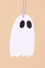 A white ghost-shaped hanging ornament with two black oval eyes and a purple string at the top, shown against a solid beige background.