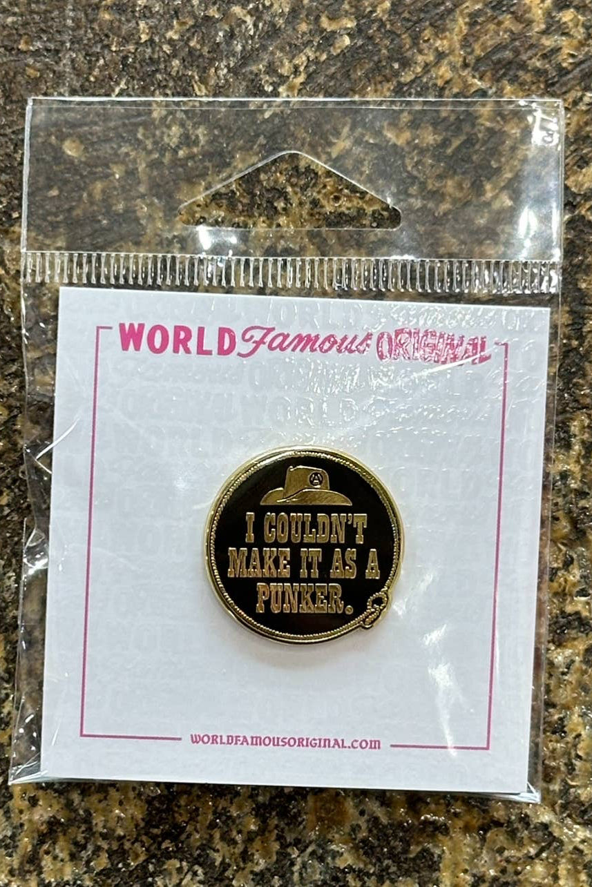 A round enamel lapel pin with a gold-tone finish and a small hat icon above the text 'I COULDN'T MAKE IT AS A PUNKS.' is shown in its clear plastic packaging on a white backing card, placed on a mottled brown and black stone surface.