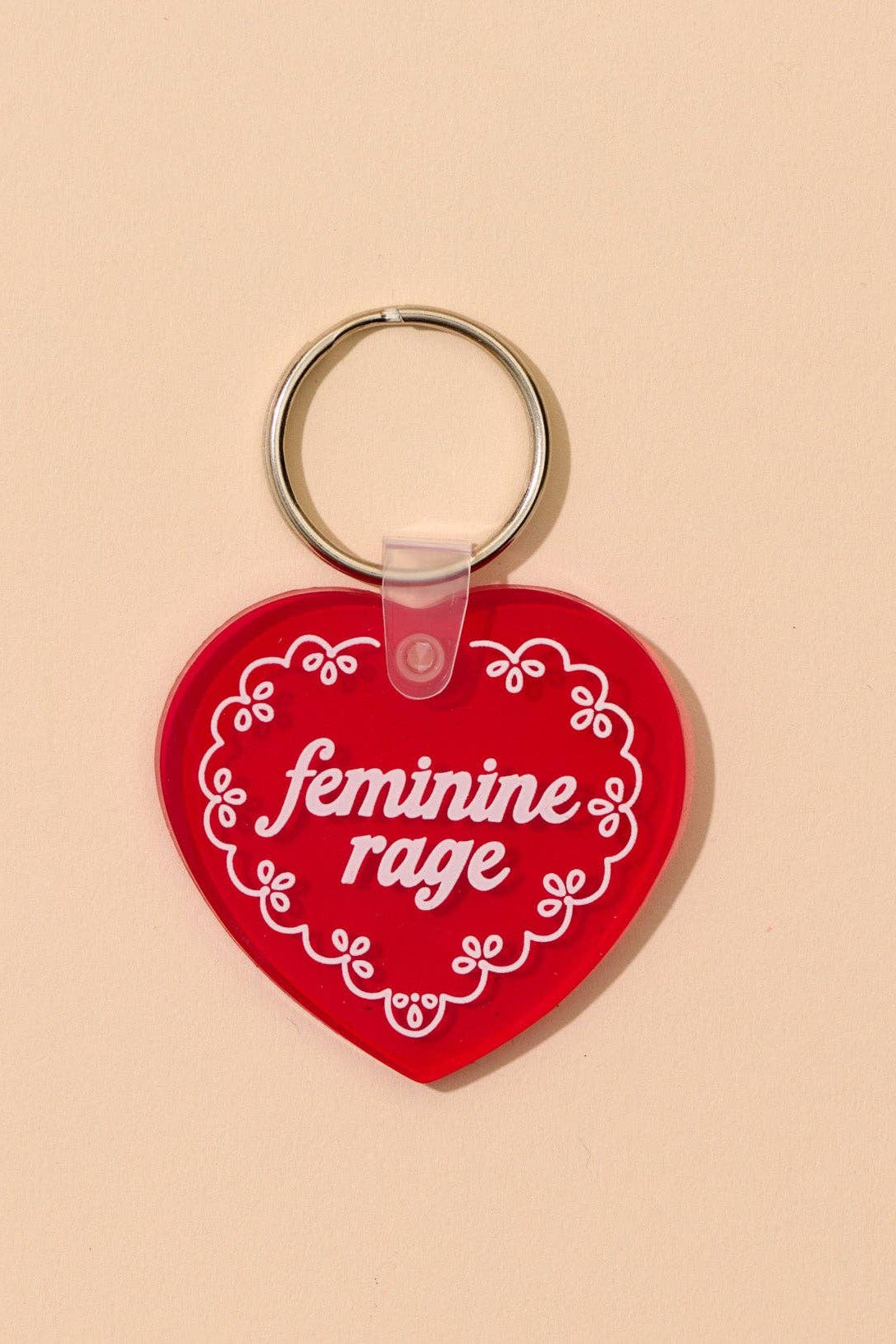 Red heart-shaped keychain with 'feminine rage' text on a beige background