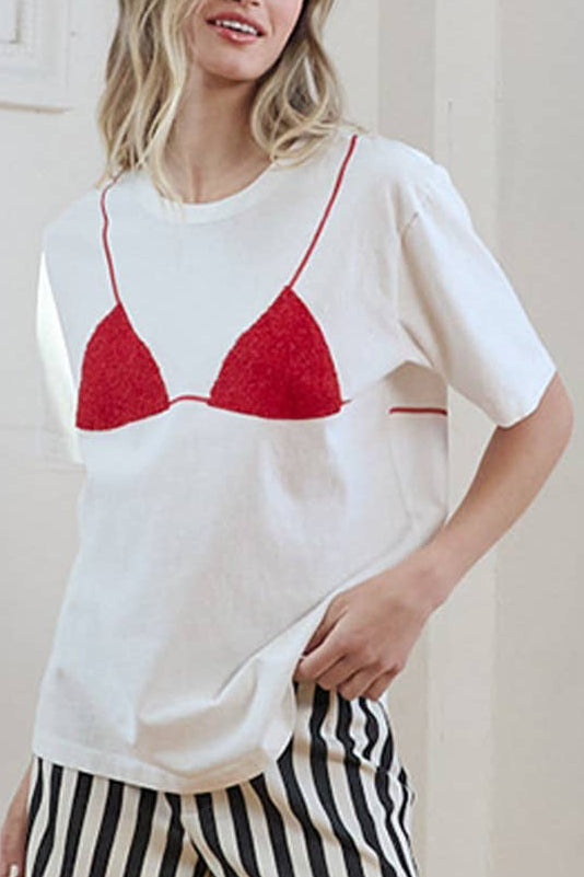 Woman wearing a white t-shirt with a red bralette and black and white striped shorts in a room.