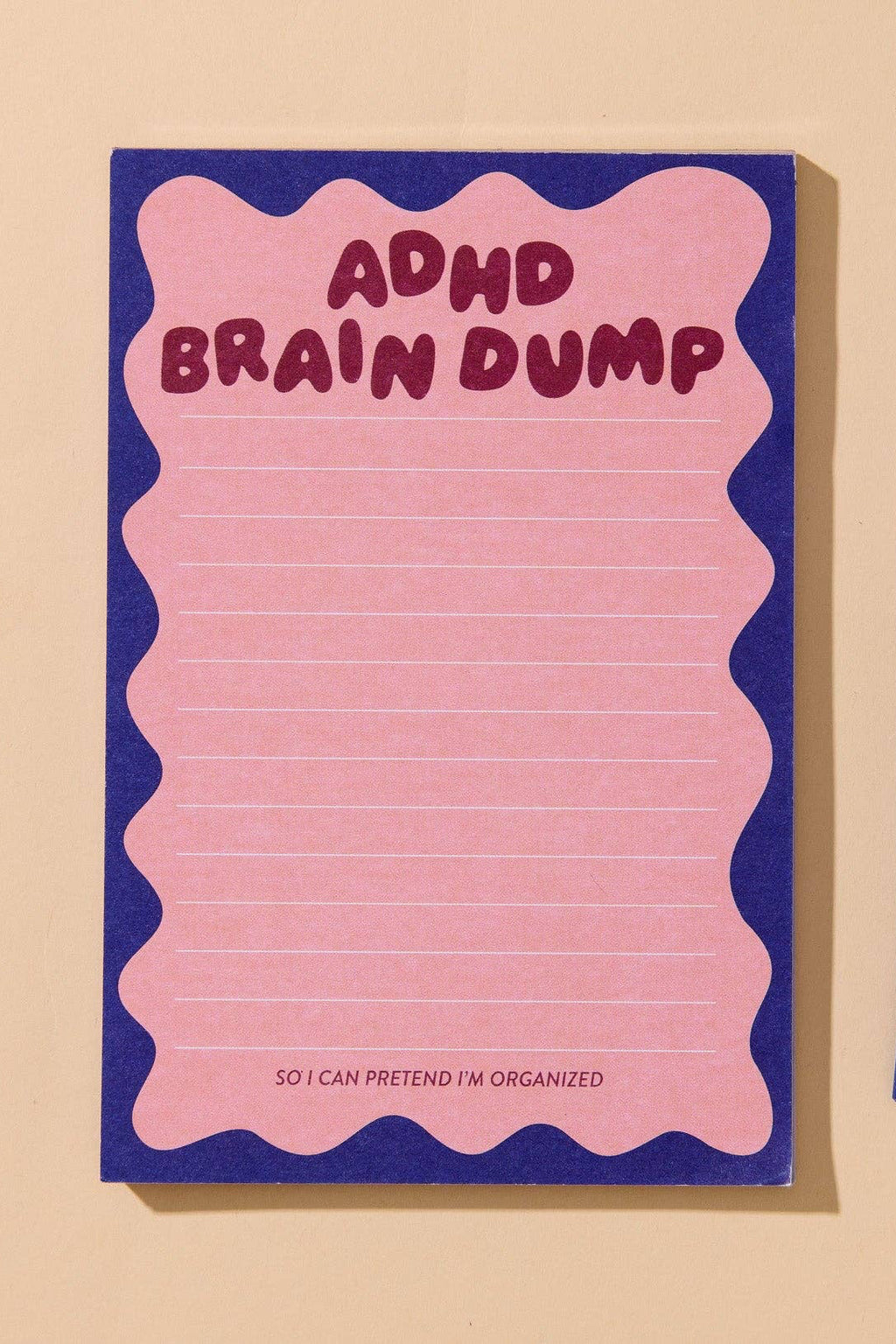 A pink and navy blue notepad with the text 'ADHD BRAIN DUMP' on the front, shown next to a blue and lavender pen on a beige background.