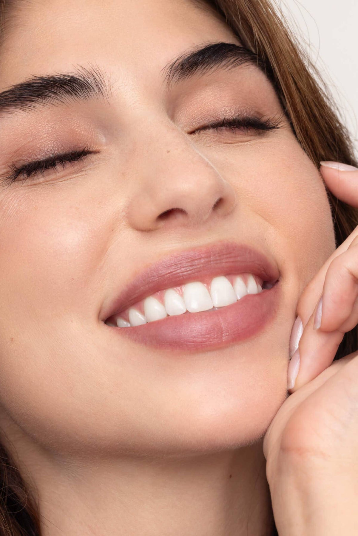 Close-up of a woman with a soft smile and hand on her cheek against a neutral background